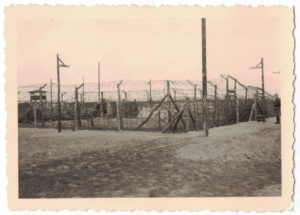 Ein mit Stacheldraht umzäuntes, von Wachttürmen flankiertes Gelände ist zu sehen. Hinten rechts steht ein Soldat mit Gewehr.