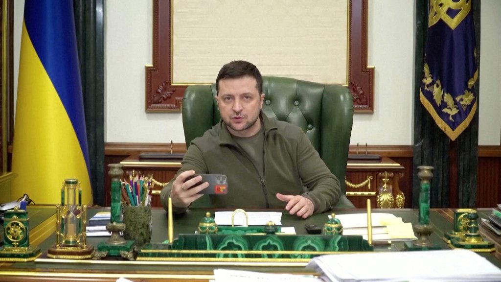 Der Präsident der Ukraine Wolodymyr Selenskyj sitzt an seinem Schreibtisch und hält eine Ansprache an seine Bevölkerung. Dabei filmt er sich selbst mit dem Handy.