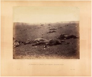 Tote Männer auf dem Rücken auf einem Feld; im Hintergrund einige Personen zu Pferde oder stehend, schwarz-weiß.
