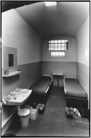 Kleine Zelle mit zwei Betten, einem Waschbecken, einem kleinen Spiegel, einem Tisch und einem vergitterten Fenster.