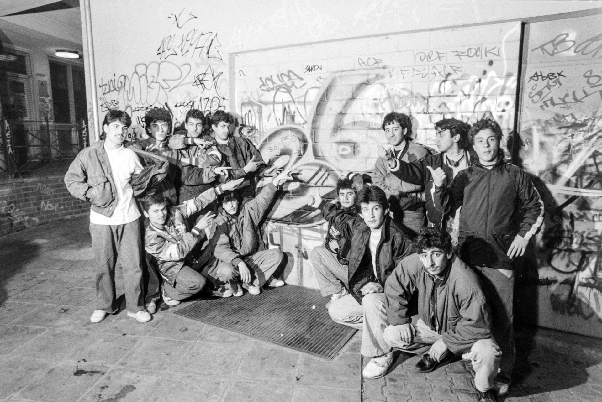 Zwölf junge Männer vor einer Wand voller Graffiti, die auf die Wand zeigen und in die Kamera schauen.