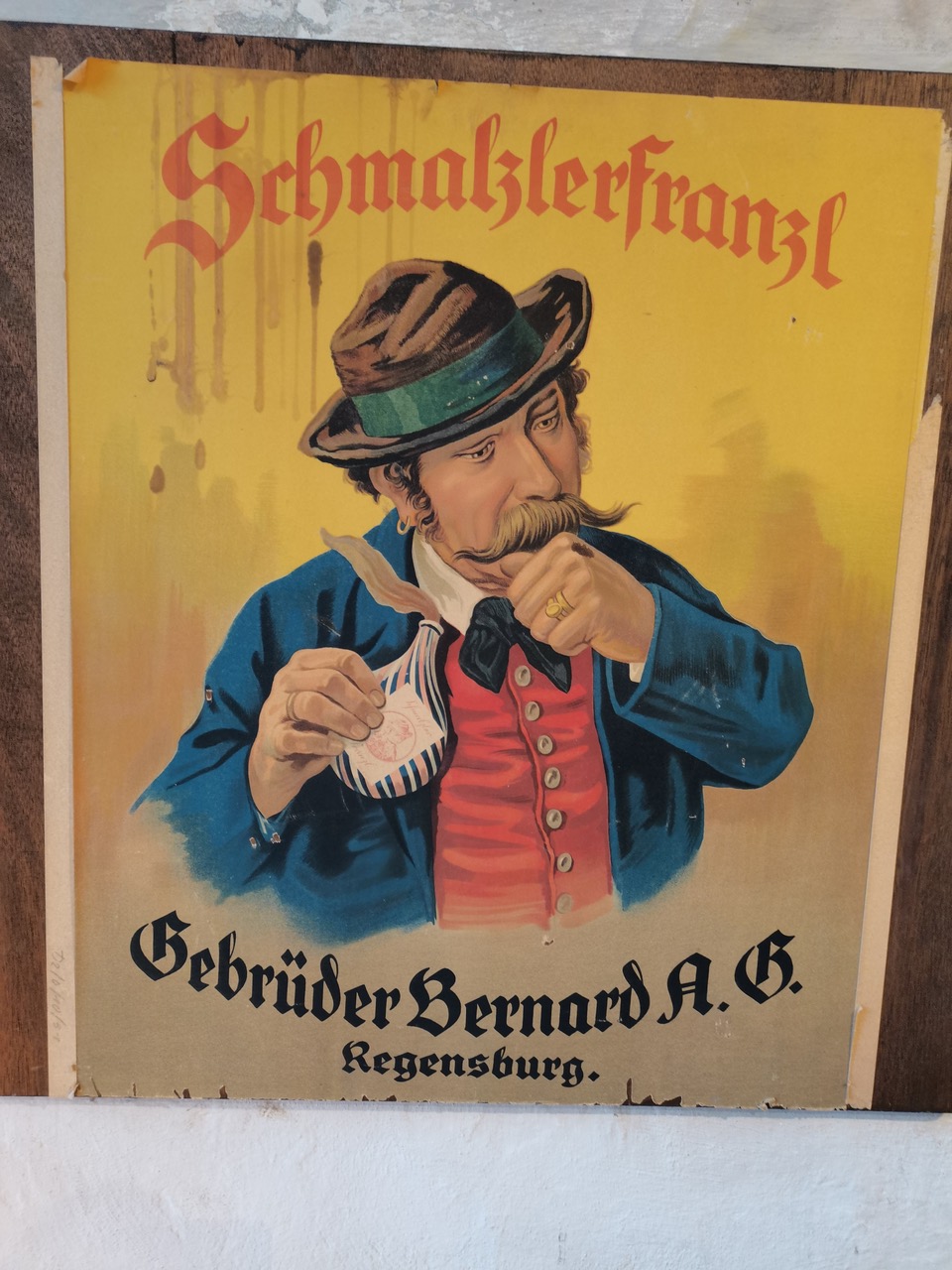 Ein Schnupftabak-Werbeplakat „SchmalzlerFranzl“, auf dem ein Mann mit Schnauzbart, Hut und Janker zu sehen ist, der den Tabak schnupft; darunter der Schriftzug „Gebrüder Bernard A.G. Regensburg“