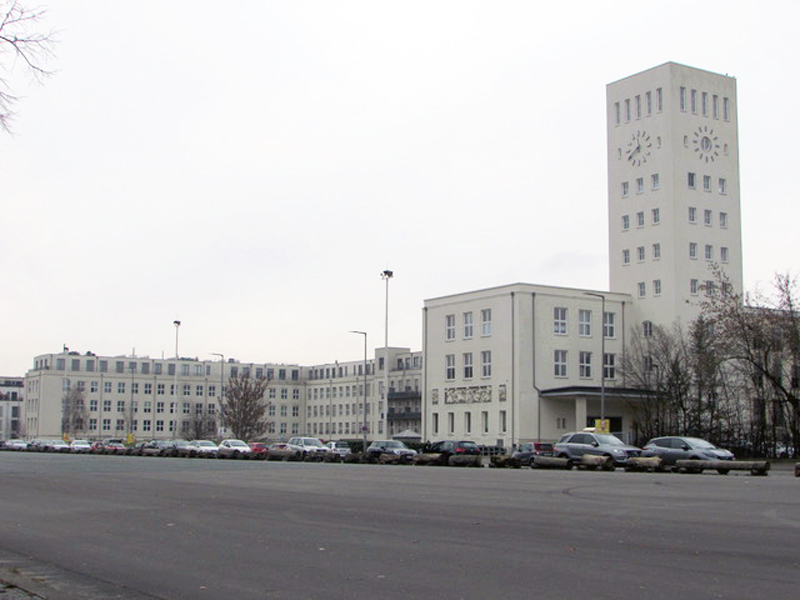 Blick von der Straße auf ein großes, rechtwinkliges, weiß verputztes Gebäude mit Uhrenturm