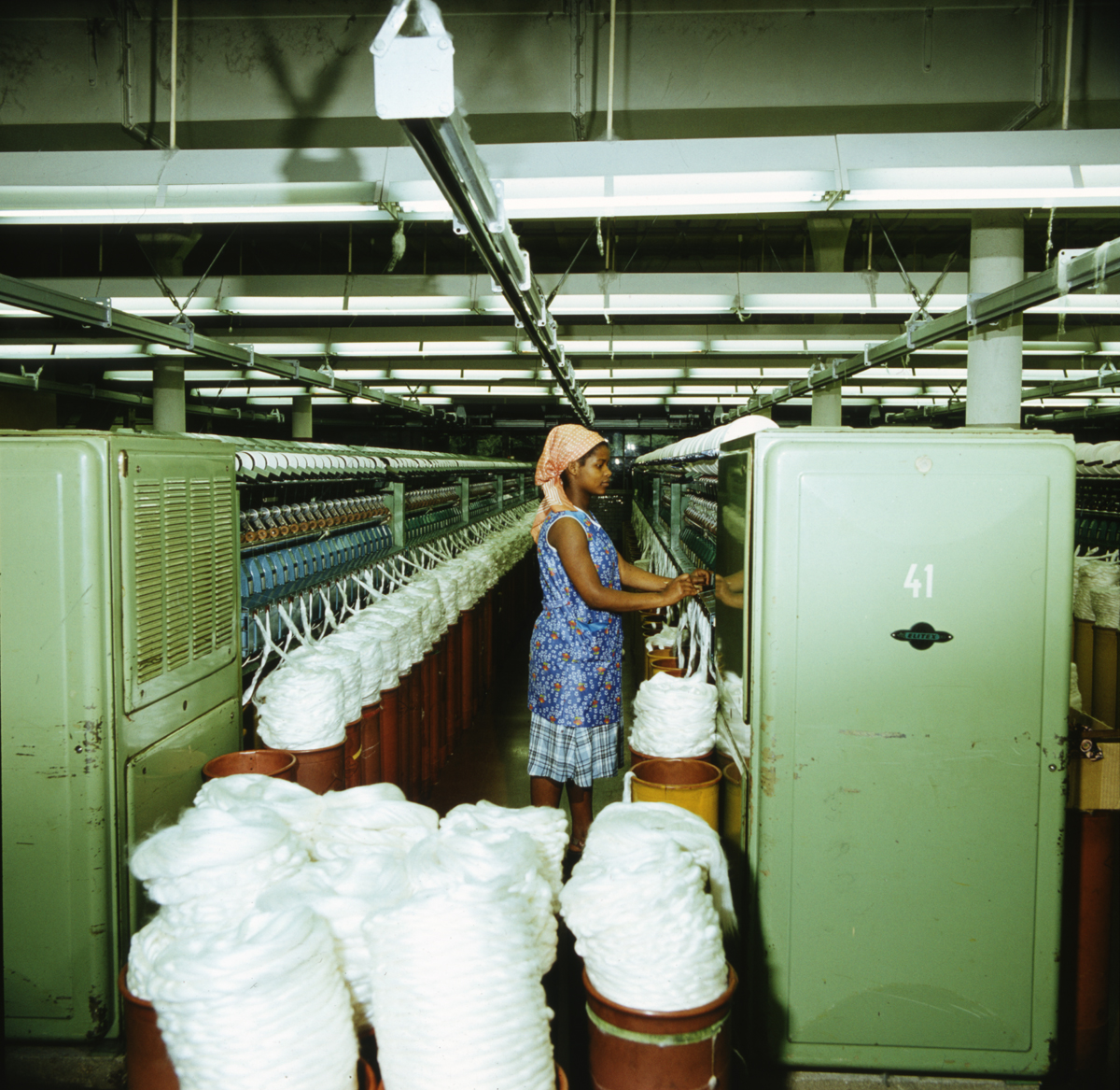 Eine Frau in einem Kittel und mit Kopftuch, die in einer Fabrik für Baumwollspinnerei arbeitet; vor ihr und hinter ihr ist die maschinell verarbeitete Baumwolle zu sehen.