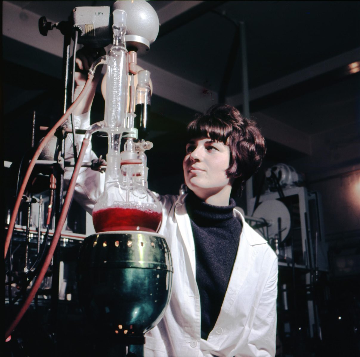 Eine Frau in einem weißen Kittel steht in einem Forschungslabor neben einem Glasgefäß, das mit roter Flüssigkeit gefüllt ist.