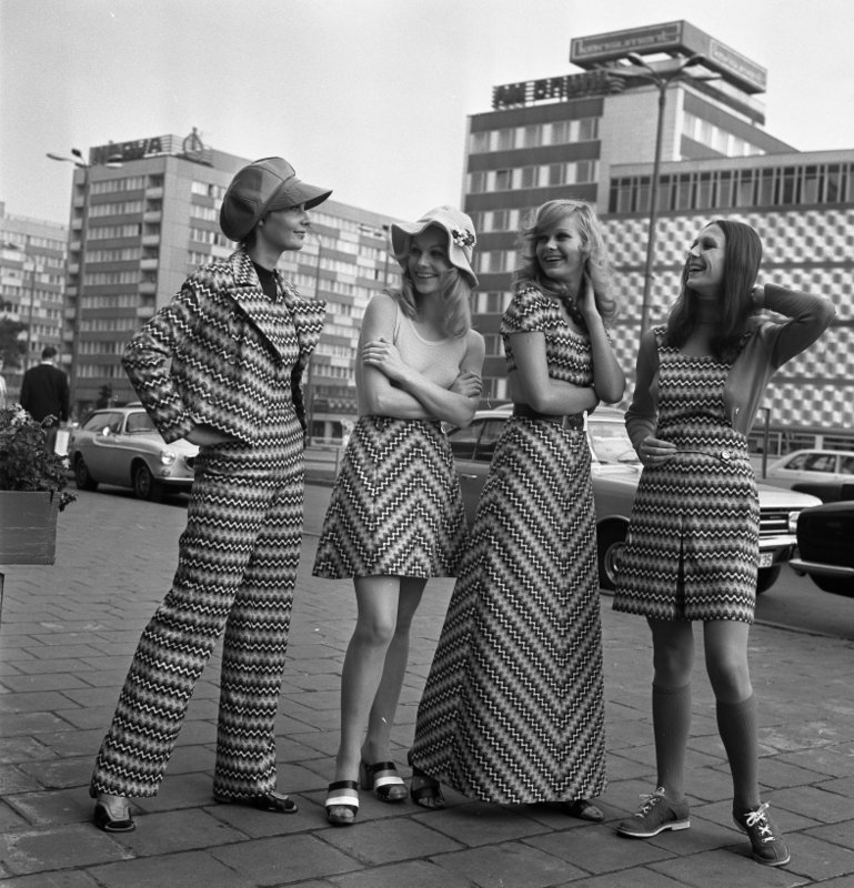 Vier junge weibliche Fotomodelle stehen nebeneinander auf einem Platz und stellen ein neues, wild gestreiftes Textilgewebe vor, aus dem ihre Kleidung – Rock, Kleider, Hosenanzug – gefertigt wurde.