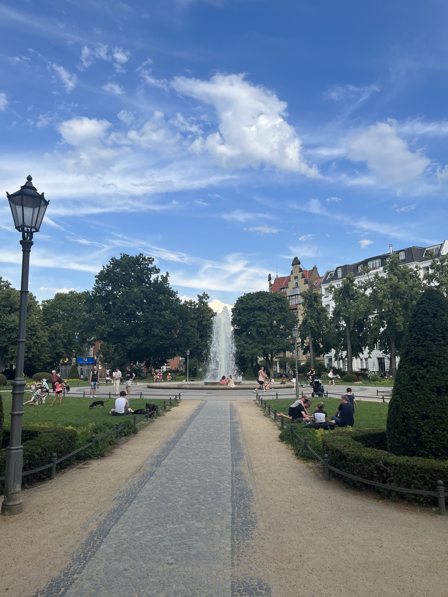 Blick in einen städtischen Park: in der Mitte ein Springbrunnen mit Fontäne