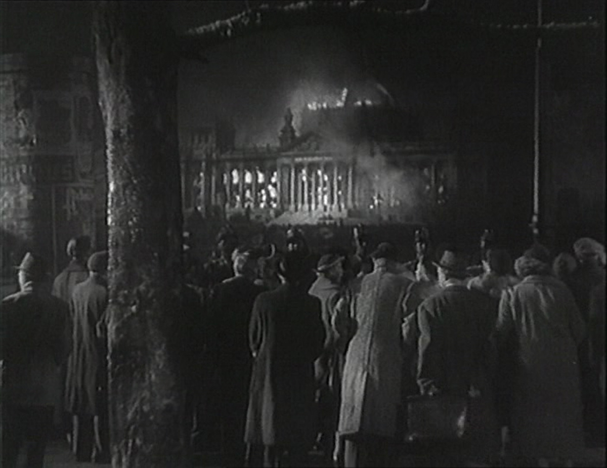 Screenshot aus dem Film „Der Teufelskreis“, das ein in Flammen stehendes Gebäude in Schwarz-Weiß zeigt, davor eine Gruppe von Passant:innen.