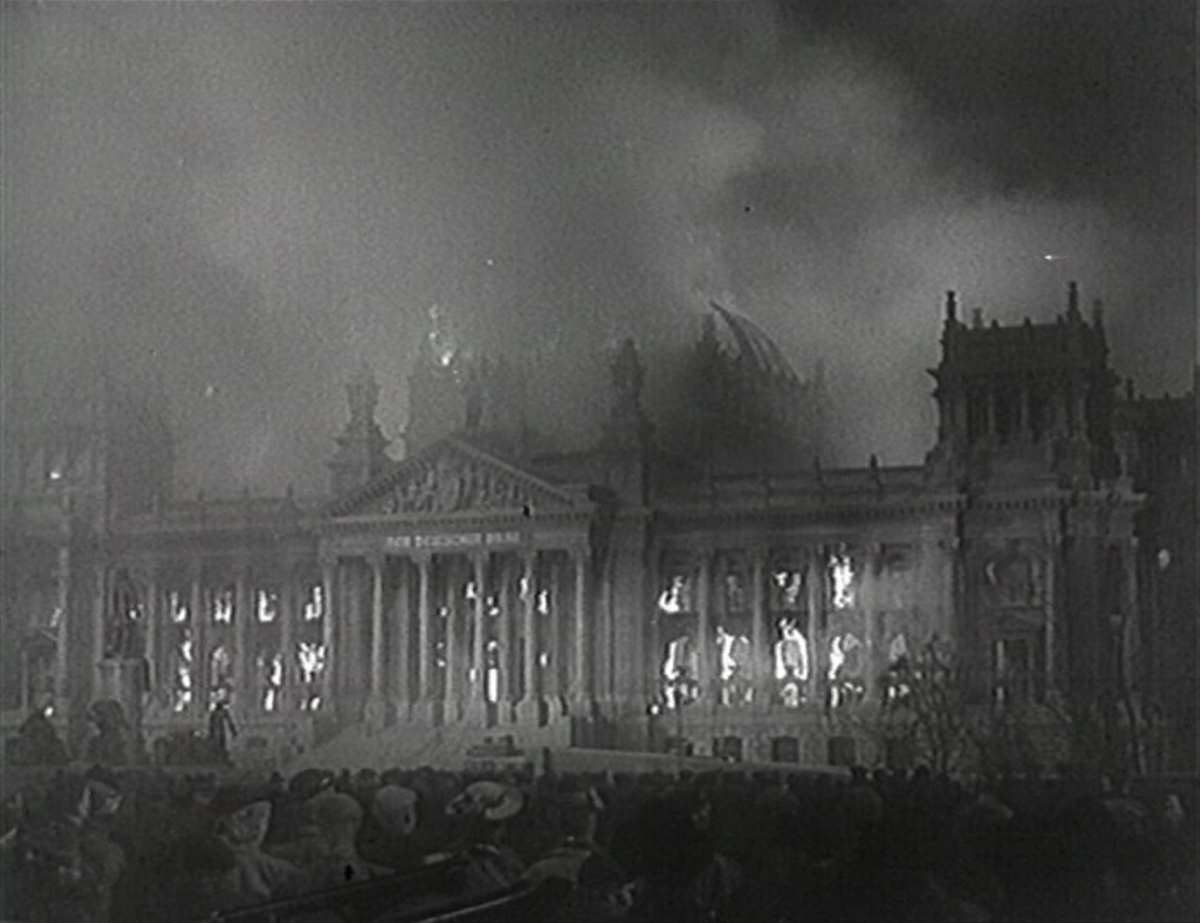 Screenshot aus dem Film „Der Teufelskreis“, das ein ein in Flammen stehendes Gebäude in Schwarz-Weiß zeigt.