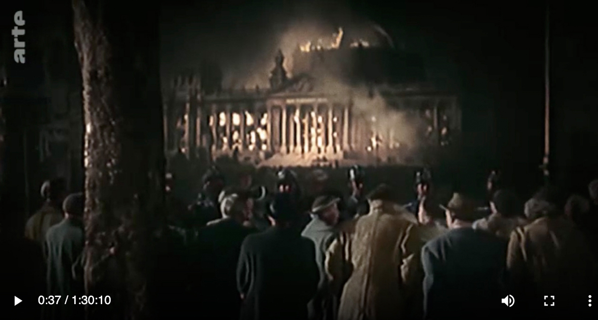 Screenshot Arte, das ein in Flammen stehendes koloriertes Gebäude zeigt, davor eine Gruppe von Passant:innen.