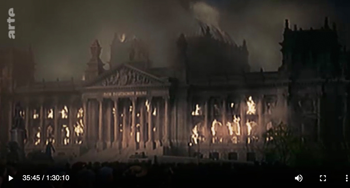 Screenshot Arte, das ein in Flammen stehendes koloriertes Gebäude zeigt.