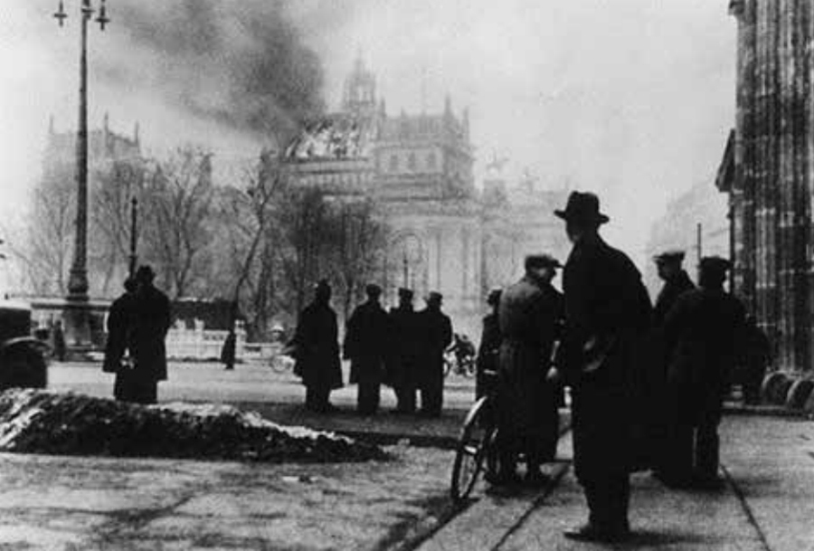 Schwarz-Weiß-Fotografie: Mehrere Menschen stehen alleine und in Gruppen auf der Straße und blicken auf ein Gebäude vor ihnen. Aus der Kuppel steigt Qualm empor, auch Flammen sind zu erkennen.