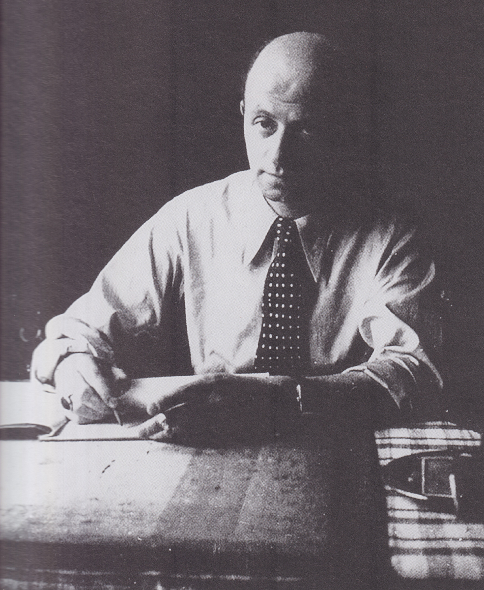Foto eines Mannes in Hemd und mit Schlips, der an einem Holztisch sitzt und etwas auf Papier aufschreibt.