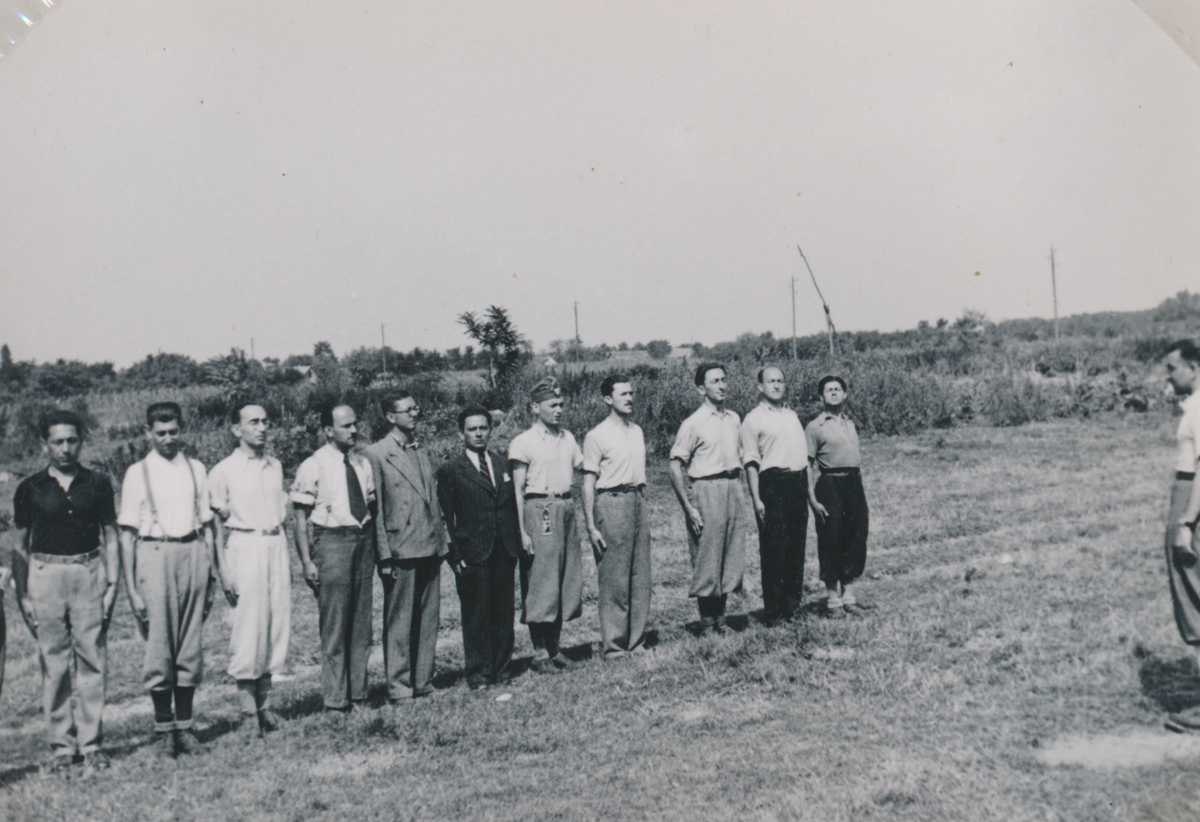 Bild von elf Männern, die in ganz unterschiedlicher Kleidung (Arbeitskleidung und im Anzug) auf einem Feld in einer Reihe aufgestellt stehen
