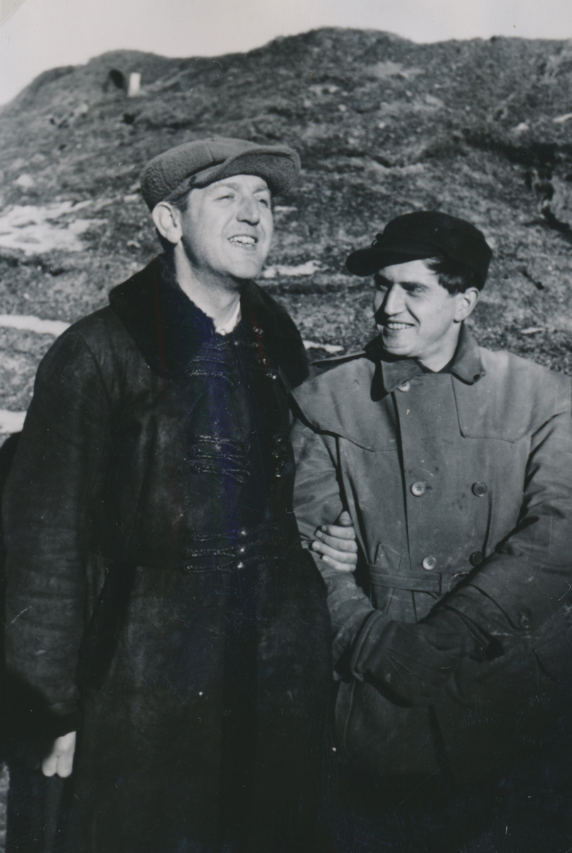 Zwei Männer stehen eng nebeneinander, der eine hält den Arm des anderen; im Hintergrund ein Hügel.
