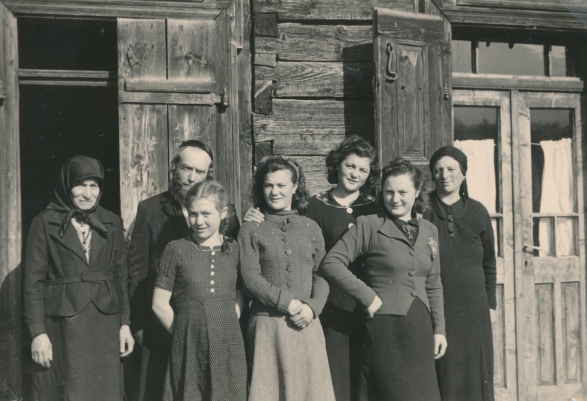 Sieben Personen (sechs Frauen und ein Mann) stehen vor einem Holzhaus.