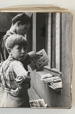Foto eines Jungen, der in die Kamera blickt; neben ihm ein Mann, der in Zeitungen blättert, die auf einer Mauer liegen.