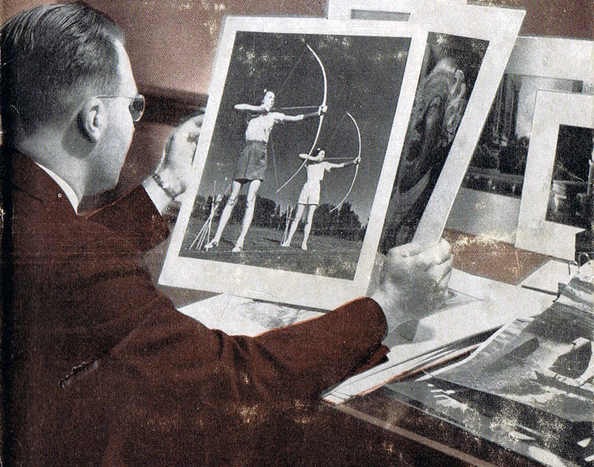 Fotografie eines Mannes, von seitlich hinter ihm fotografiert, an seinem Schreibtisch, der mehrere Fotografien betrachtet; eine zeigt zwei Personen, die mit dem Bogen schießen.