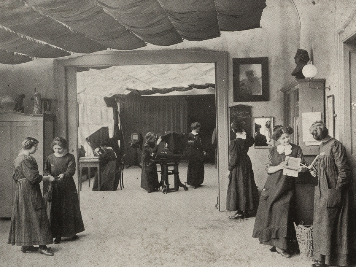 In einem großen zweiteiligen Zimmer sind mehrere junge Frauen in langen Kleidern zu sehen, die verschiedenen Tätigkeiten mit der Kamera nachgehen.