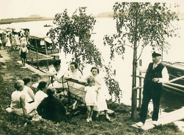 Eine Familie sitzt auf einer Bank bzw. auf dem Rasen an einem Fluss.