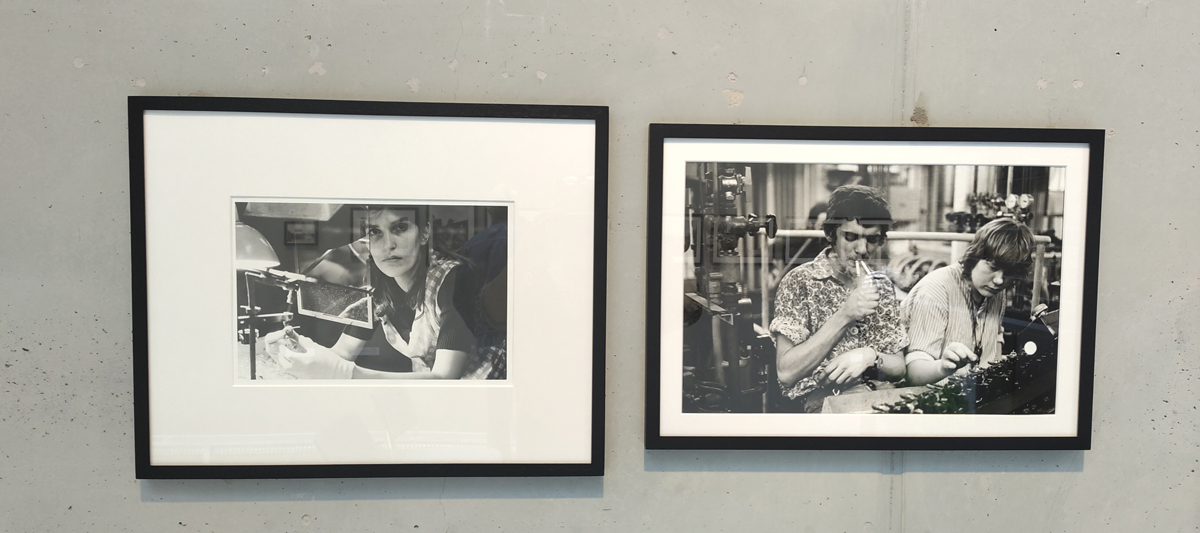 Zwei Schwarzweiß-Fotos hängen an einer Wand: links: eine Frau im Kittel arbeitet mit Lampen; rechts: ein Mann, mit zwei Zigaretten im Mund, und eine Frau an einem Tisch in einer Industriehalle
