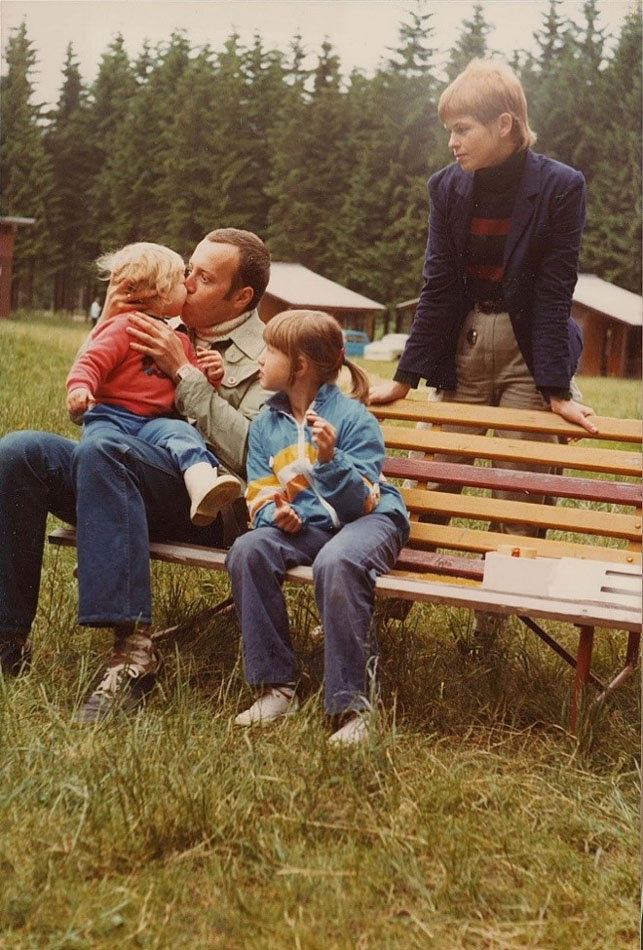 Ein Mann auf einer Bank mit drei Kindern um ihn herum, dahinter Tannen