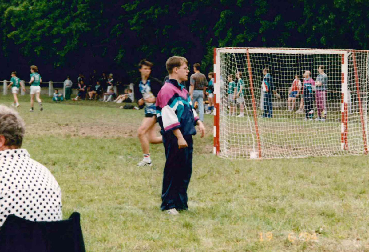 Spielende Jungs und Zuschauer auf einem Rasenplatz, im Vordergrund ein Tor und ein Junge in Trainingsjacke- und Hose