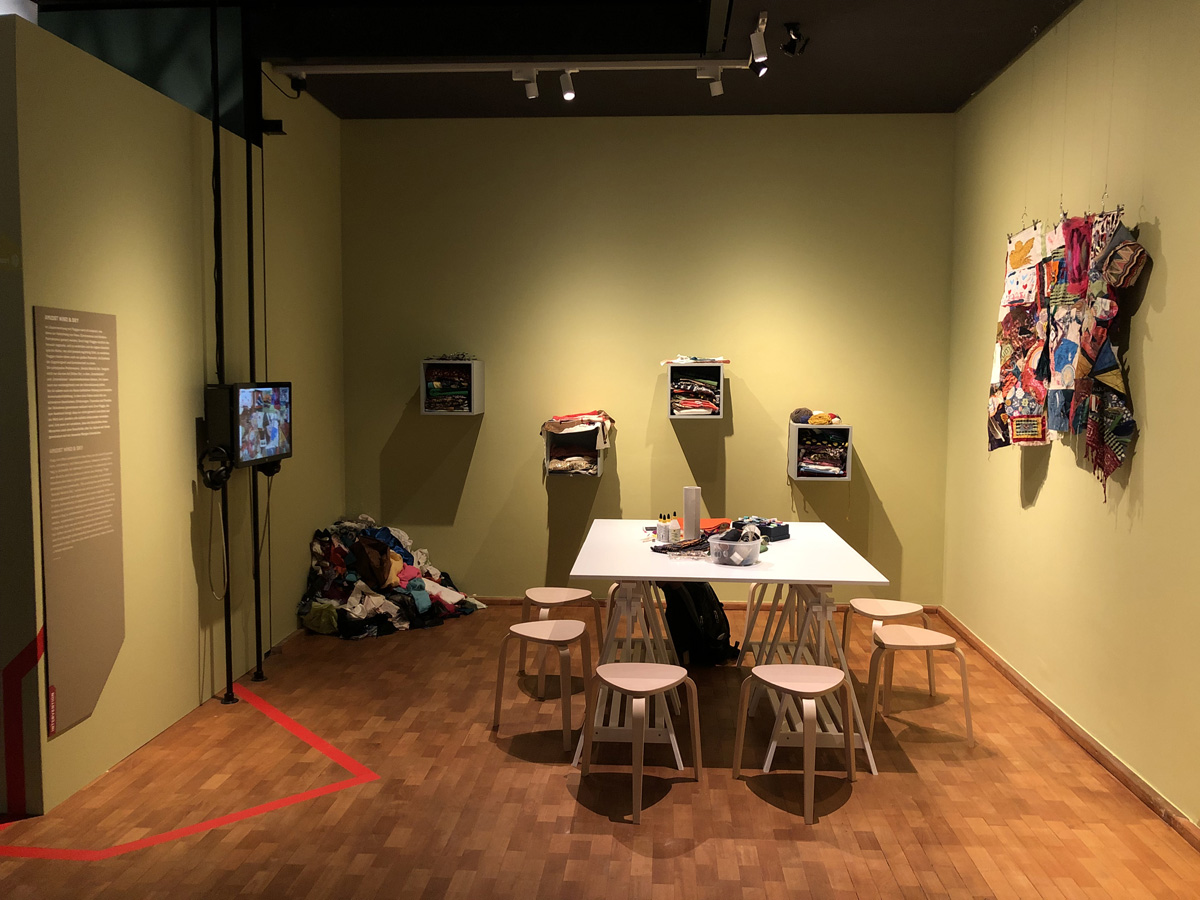 Raum mit Tisch und Stühlen; an der Wand Stoffe, ein Bildschirm und eine Texttafel