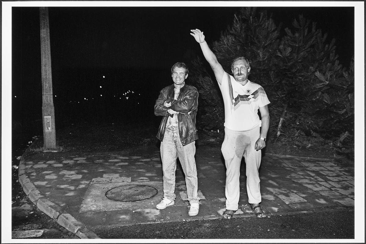 Schwarzweiß-Foto eines Mannes, der im weißen Fußballshirt und in Jogginghose den rechten Arm hebt; neben ihm ein lachender Mann mit verschränkten Armen.