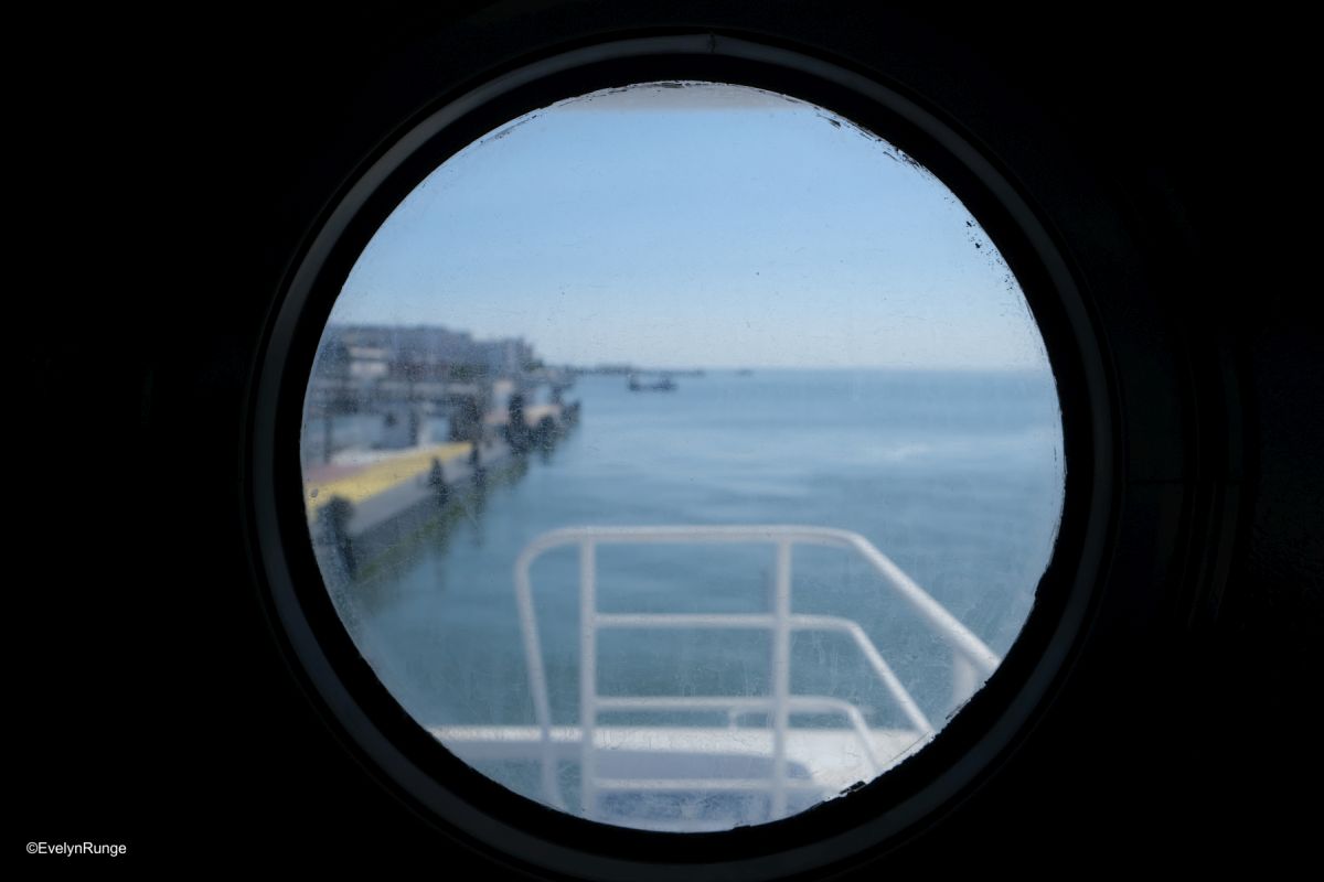 Ausblick aus einem Bullauge: rechts Meer, links Hafenindustrie