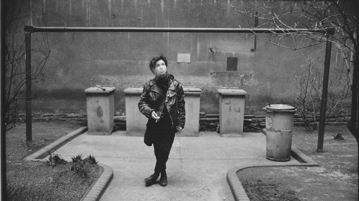 Schwarzweiß-Foto einer jungen Frau in einer Lederjacke in einem Hinterhof vor Mülltonnen