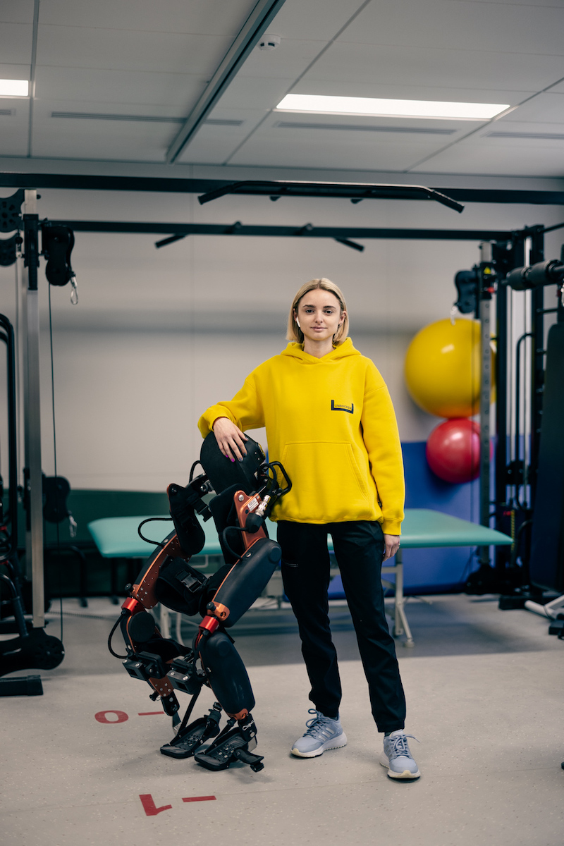 Farbfoto einer jungen Frau in einem Sportstudio mit einem Gehroboter neben sich