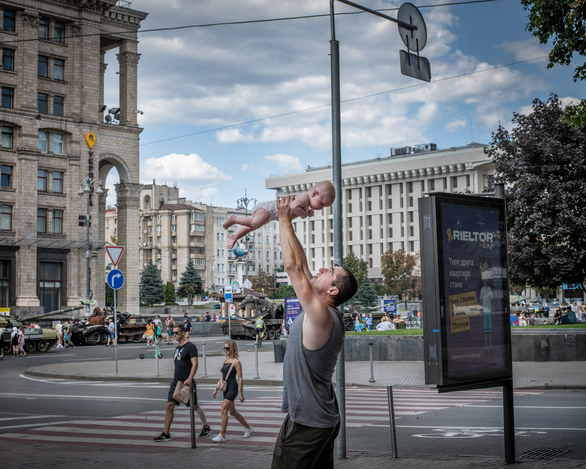 Farbfoto eines Mannes in einer Straße, der ein Kind in die Luft hebt; hinter ihm ein Platz mit zerstörten Panzern.