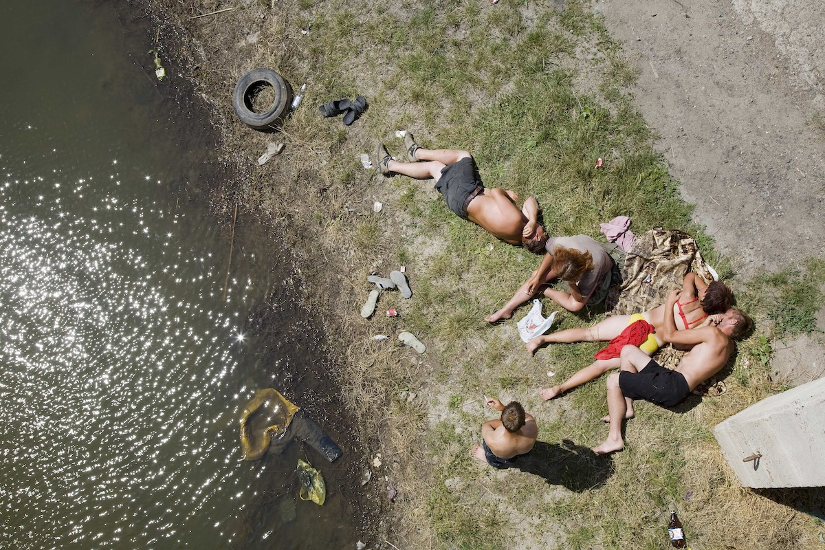 Farbfoto: Blick von oben auf fünf Personen, die sich in Badekleidung am verschmutzten Ufer eines Flusses aufhalten.