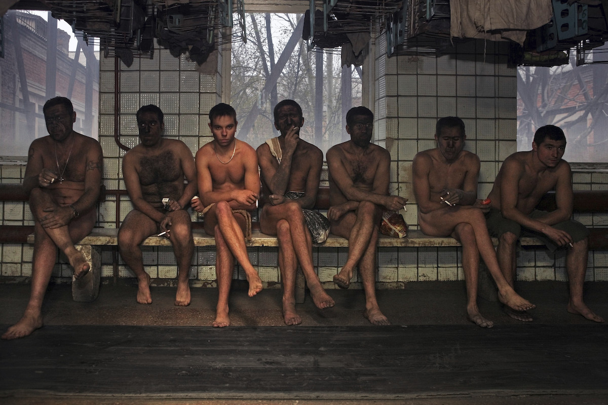 Farbfoto von sieben nackten Männern in einem gekachelten Raum, die dort auf einer Bank sitzen.