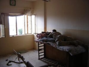 Farbfotografie eines Mannes, der mit Gewehr auf dem Bett in einem Zimmer liegt und durch das Fenster nach draußen zielt.