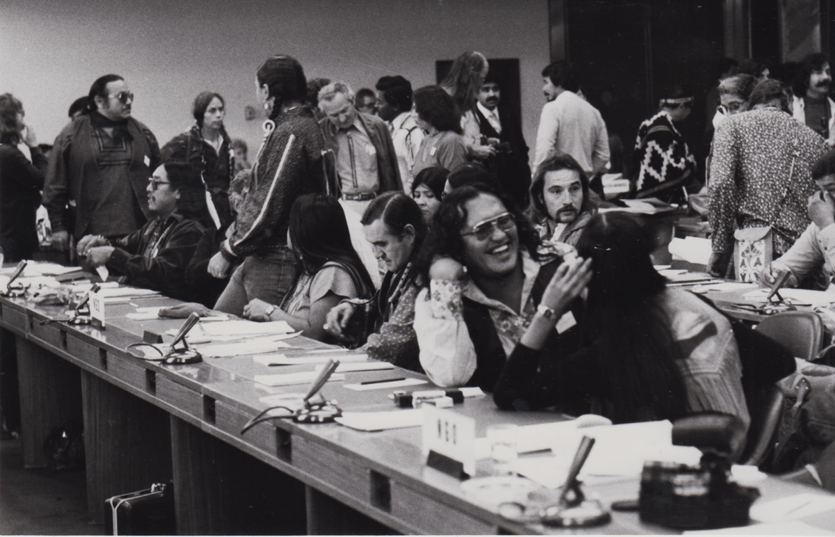 Schwarzweißfoto von sitzenden und stehenden Menschen in einem Raum mit Konferenztischen
