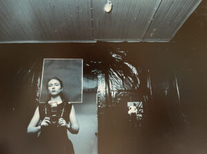 Schwarzweißfoto einer Person mit einer Kamera in den Händen und einer Art Plakat in Höhe des Kopfes