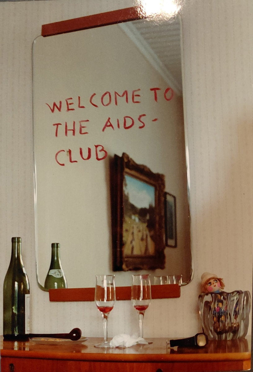 Farbfoto von einer leeren Flasche und Gläsern, darüber ein Spiegel, auf dem steht: "Welcome to the AIDS-Club"