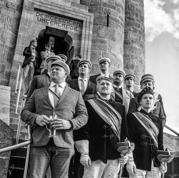 Schwarzweißfotografie von Männern mit Schärpen und Mützen vor einem Backsteingebäude