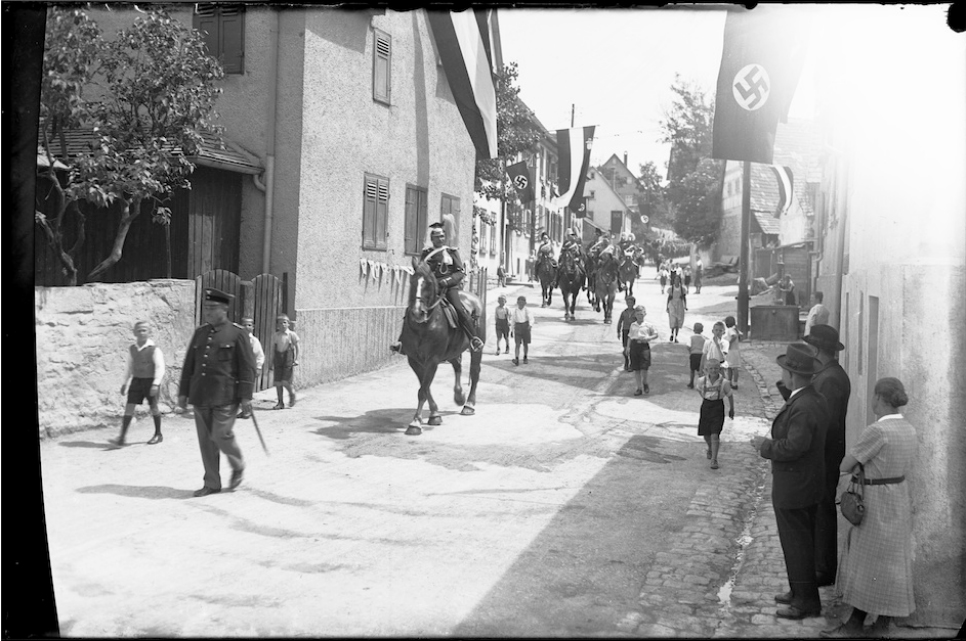 Schwarzweißfoto von Männern auf Pferden auf einer dörflichen Straße mit wenigen Zuschauern