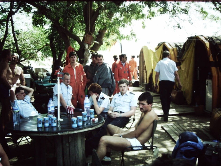 Farbfotografie von Männern an einem Tisch unter Bäumen; daneben Zelte