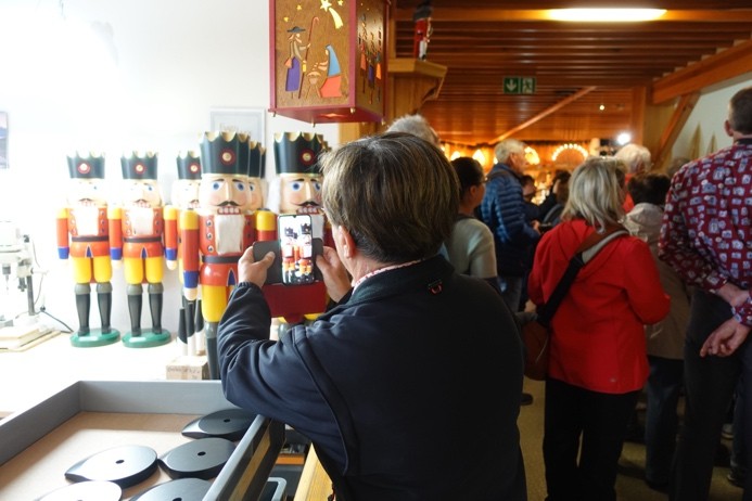 Farbfoto von vielen Menschen in einem Raum; im Vordergrund eine Person, die mit dem Handy hölzerne Nußknacker fotografiert.