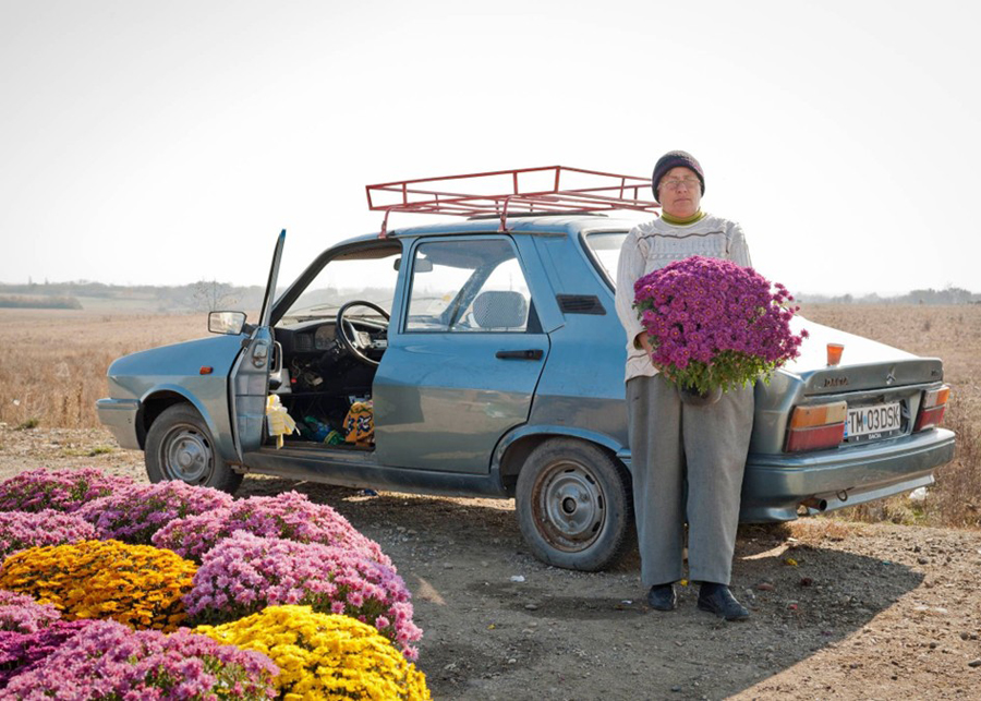 Farbfoto einer Frau mit Blumen in der Hand vor einem blauen Auto, daneben weitere Blumen
