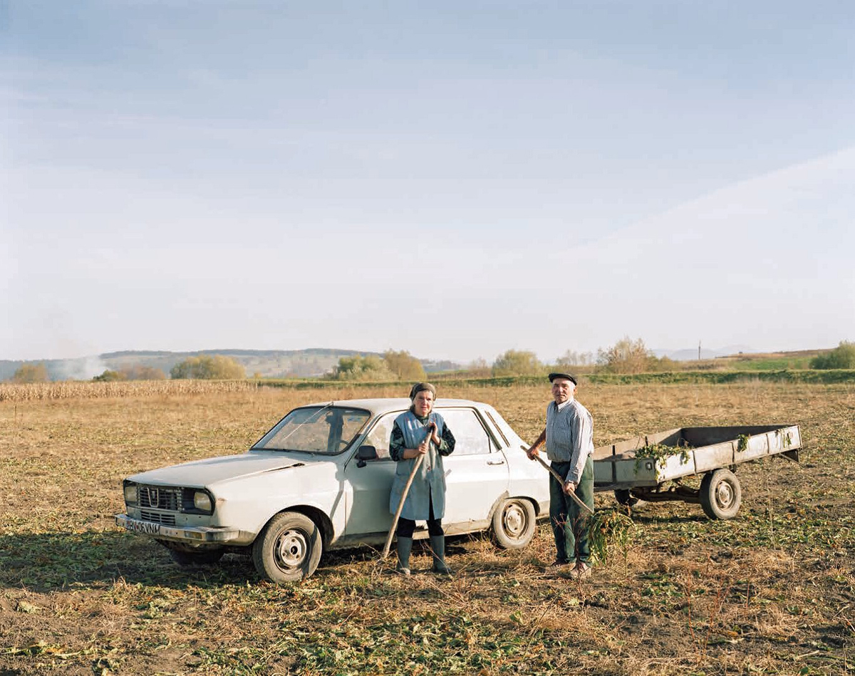 Farbfoto von zwei Personen mit Hacken auf einem Feld, neben ihnen ein weißes Auto mit Hänger