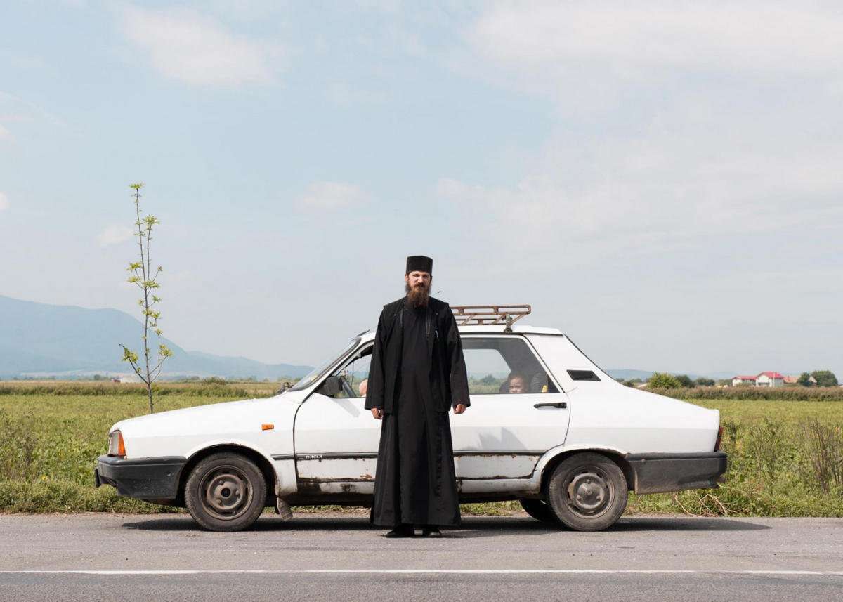 Farbfoto eines Geistlichen vor einem weißen Auto auf einer einsamen Straße