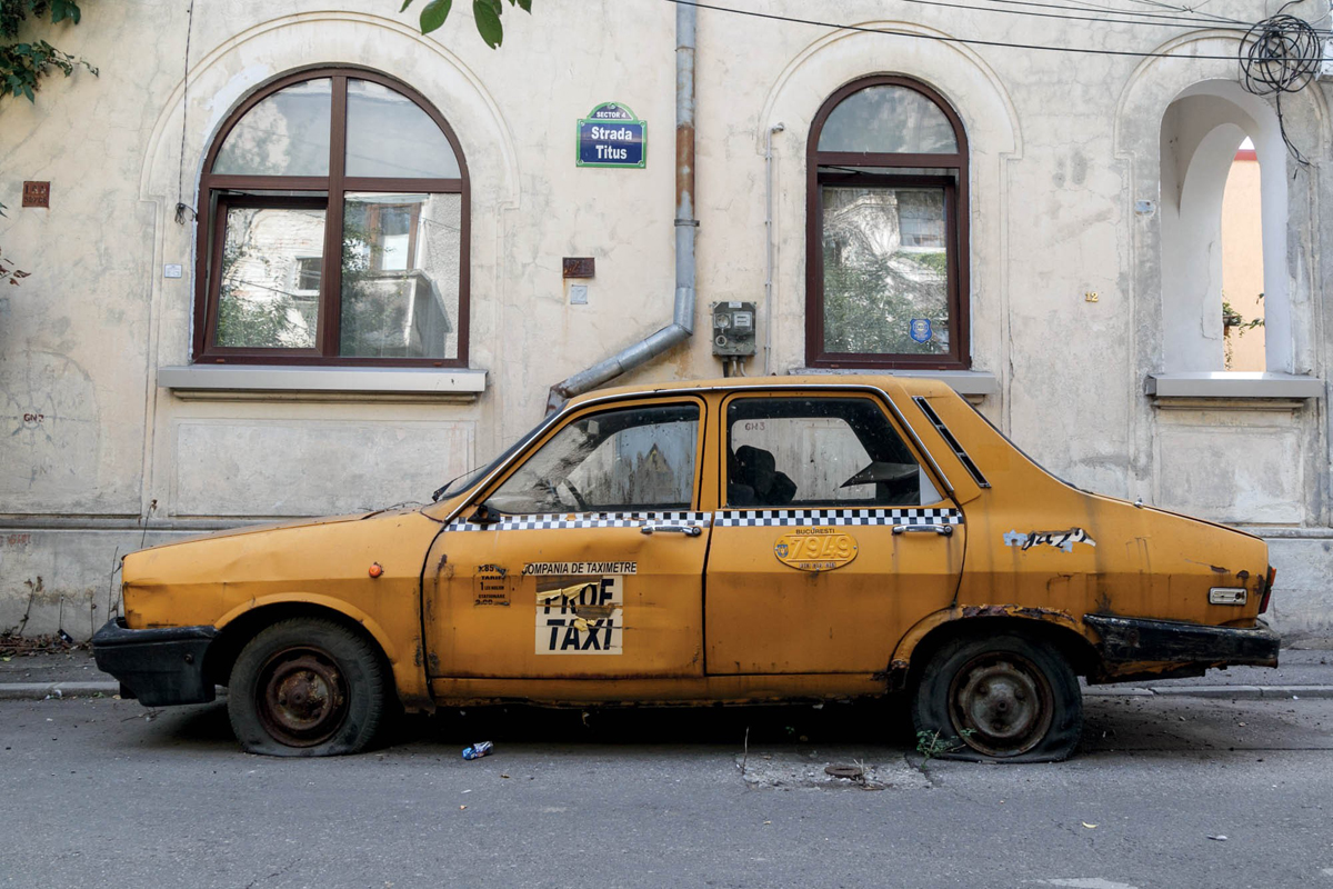 Farbfoto eines gelben, alten und verbeulten Autos mit platten Reifen und dem Aufdruck Taxi in einer Straße