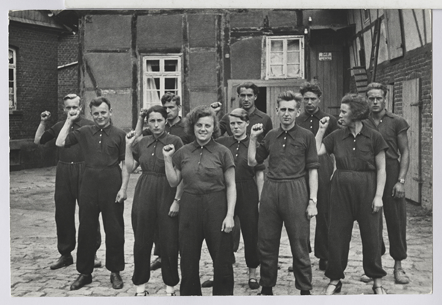 Schwarzweißfoto von jungen Männern und Frauen stehend mit erhobener Faust vor einem Bauernhof.