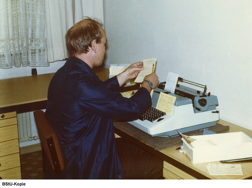 Farbfoto eines Mannes an einer Schreibmaschine mit Aktenmaterial