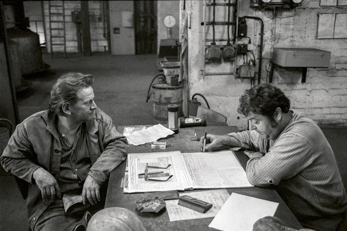 Schwarzweißfoto von zwei Männern in Arbeitskleidung an einem Tisch sitzend in einer Fabrik